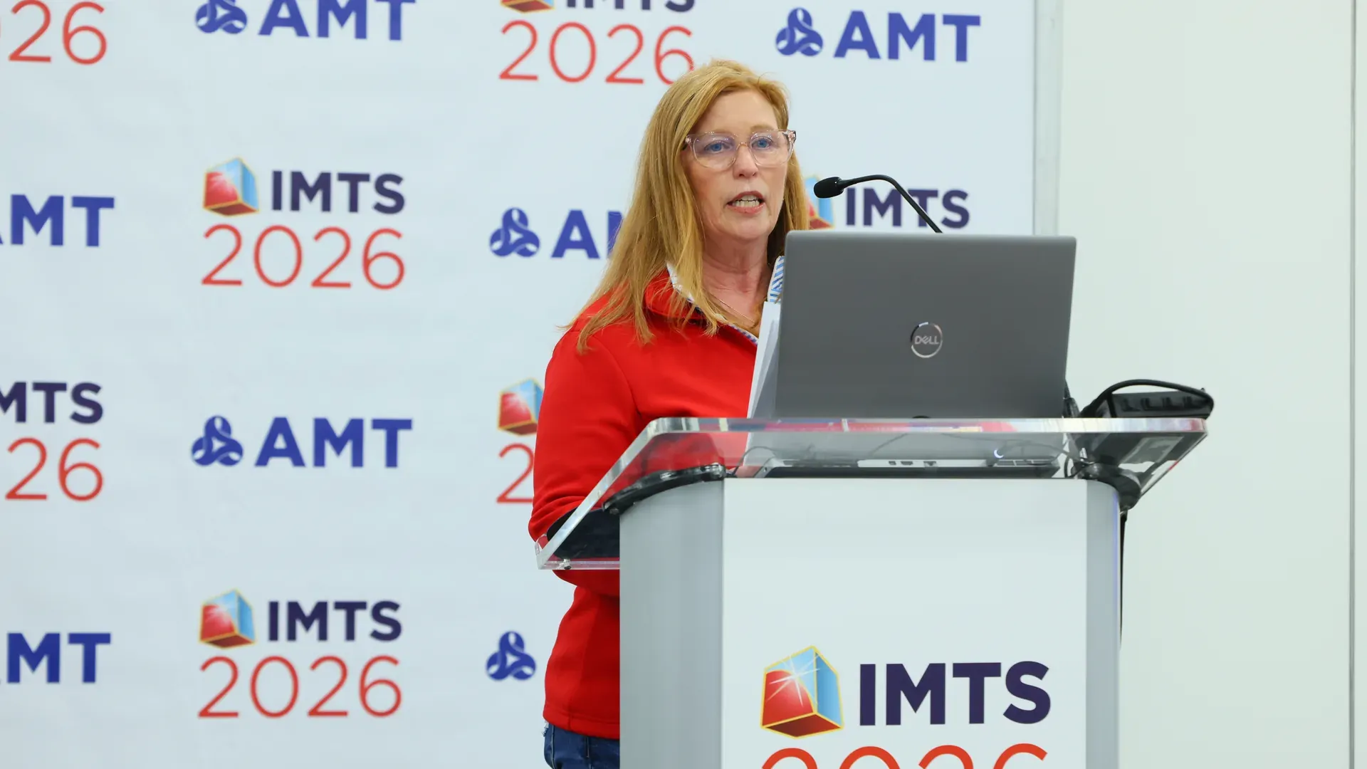 Bonnie Gurney of AMT speaking at a lectern in front of an IMTS 2026 and AMT branded backdrop